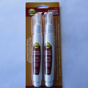 May include: Two white tubes of Aleene's Fabric Fusion permanent fabric adhesive. The tubes are labeled with the product name and instructions for use. The packaging is gold with the words "Fabric Fusion" in black.