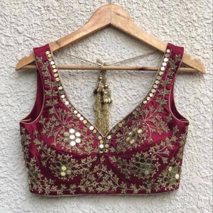 Könnte beinhalten: Eine ärmellose Bluse in Bordeauxrot mit einem tiefen V-Ausschnitt. Die Bluse ist mit goldener Stickerei und Spiegelarbeiten in einem Blumenmuster verziert.