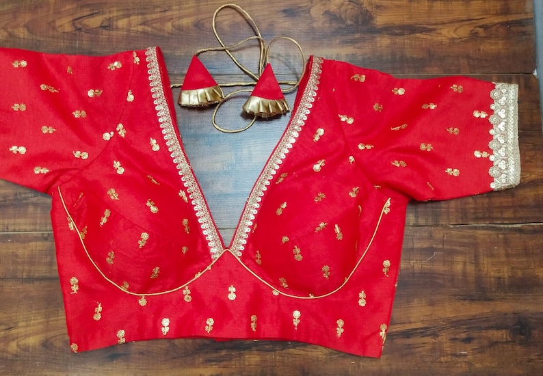 Pu&ograve; includere: Una blusa rossa ricamata con bordi dorati e una profonda scollatura a V. La blusa ha maniche corte e un corpetto aderente.