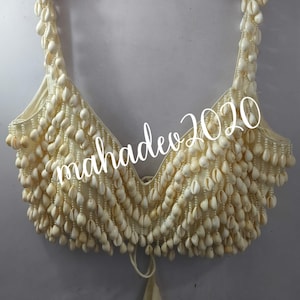 May include: A white crop top made of small white seashells. The top has a V-neck and thin straps. The top is tied at the back with a white string.