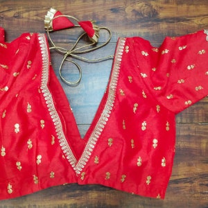 Pu&ograve; includere: Una blusa di seta rossa con ricami dorati e un bordo in pizzo argentato. La blusa ha una scollatura a V profonda e maniche corte.