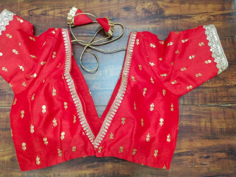 Pu&ograve; includere: Una blusa di seta rossa con ricami dorati e un bordo in pizzo argentato. La blusa ha una scollatura a V profonda e maniche corte.