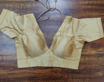 Puede incluir: Una blusa de satén dorada con escote en V y mangas cortas. La blusa tiene un ribete decorativo de lentejuelas alrededor del escote y las mangas.