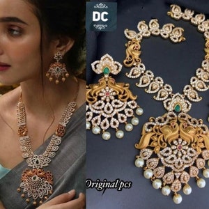 Puede incluir: Un conjunto de collar y pendientes de oro con un diseño de pavo real. El collar presenta un gran colgante con dos pavos reales enfrentados. Los pendientes son más pequeños y presentan un solo pavo real. El conjunto está adornado con piedras blancas y acentos verdes.