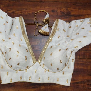 May include: White embroidered blouse with gold sequins and trim. The blouse has a deep V-neckline and short sleeves.