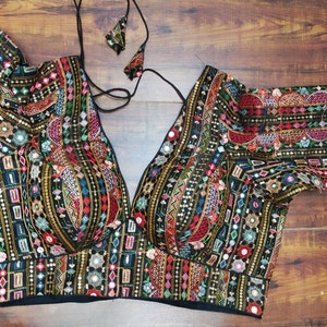 Pode incluir: Uma blusa preta bordada com decote em V profundo e mangas curtas. A blusa é decorada com padrões geométricos coloridos, lantejoulas e trabalho de espelho.