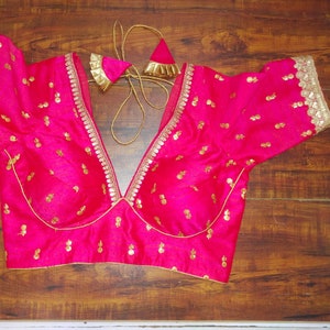 Puede incluir: Una blusa bordada en rosa y dorado con un escote en V profundo y mangas cortas. La blusa está decorada con pequeñas lentejuelas doradas y un ribete dorado.