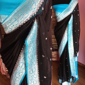 May include: A black chiffon saree with a turquoise and silver border. The saree is draped on a mannequin and the border is visible on the bottom and the pallu.