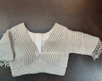 May include: A beaded, light gray blouse with a deep V-neck and three-quarter sleeves. The blouse is adorned with rows of small beads, creating a textured effect. The sleeves are finished with beaded fringe.