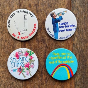 May include: Four circular anti-smoking badges. One reads "Join the majority, be a non-smoker" with a cigarette. Another says "Lungs are for life... Don't blow it!" with a person playing a trumpet. One says "Smoking Stinks" with flowers. The last says "There are no cigarettes at the end of the rainbow."
