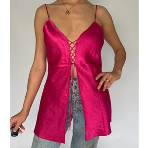 May include: A hot pink satin top with spaghetti straps and a lace-up front. The top is open down the center and worn with light wash denim jeans.