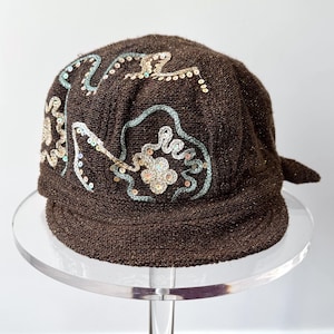 May include: A dark brown knit cap featuring a short brim and decorative embroidery. The surface is adorned with light blue and gold-toned sequins arranged in floral and swirling patterns across the front and side of the textured fabric.