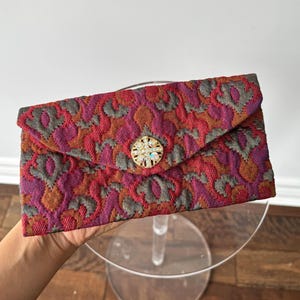 以下が含まれることがあります： 赤、紫、オレンジの色合いのカラフルな花柄が特徴のヴィンテージクラッチバッグ。 フラップクロージャーは、装飾的で丸い宝石ボタンで固定されています。 バッグは透明なスタンドで支えられています。