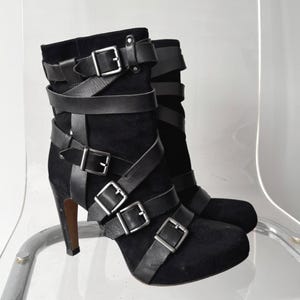 May include: Black suede ankle boots with multiple leather straps and silver-tone buckles. The boots have a high heel and a rounded toe. The straps crisscross the front and sides of the boots. The boots are displayed on a clear chair.
