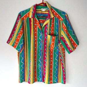 May include: A colourful short-sleeved shirt with a striped pattern. The shirt has vertical stripes in green, yellow, orange, pink, and blue, with zigzag and dotted details. It features a collared neckline and a single pocket.