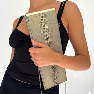 Peut inclure: Une pochette rectangulaire avec un motif peau de serpent doré et noir. Elle est dotée d'une chaîne argentée et d'un fermoir doré. La personne sur l'image porte un haut et une jupe noirs.