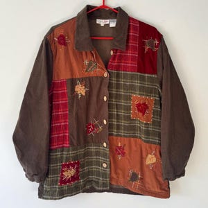 May include: A patchwork jacket with a brown corduroy collar and sleeves. The jacket features a mix of fabrics in shades of brown, red, and green, with embroidered leaf and floral accents. It is buttoned up and hanging on a red hanger.