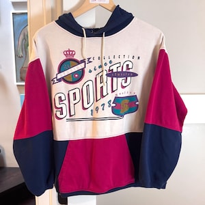 May include: A vintage-style hoodie in cream, fuchsia, and navy blue. The front features the text "SPORTS COLLECTION since 1978" in a bold, retro font. The sleeves and hood are navy blue, with fuchsia panels on the sides.