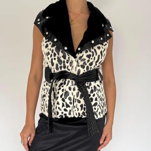 May include: A sleeveless vest with a black and white spotted pattern, a black velvet collar, and a black belt. The vest is paired with a black slip skirt with a lace hem and black boots. The outfit is a mix of textures and patterns.