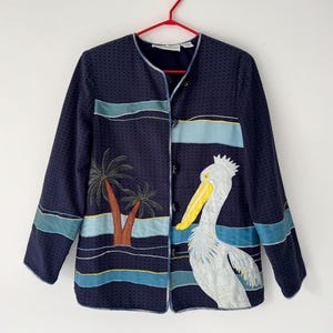 May include: A navy blue jacket with a white pelican and palm trees embroidered on the front. The jacket has a button closure and long sleeves.