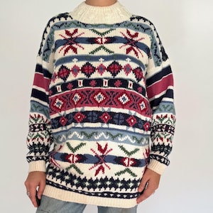 May include: Cream-colored, patterned sweater with a high neck. The sweater features a geometric design in red, blue, green, and white. The sleeves have a striped pattern in red, blue, and navy. The sweater is paired with light blue jeans.