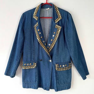 May include: A blue denim blazer adorned with gold-coloured studs and gems. The jacket features a double-breasted design with two front pockets. It is hanging on a red hanger.