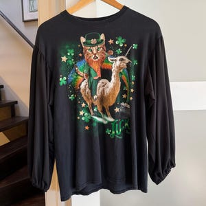 May include: Black long-sleeved top with a colourful graphic of a cat in a leprechaun hat riding a llama, with shamrocks, stars, and a rainbow. The word "LUCK" is printed in green. The shirt has a crew neck and gathered sleeves.