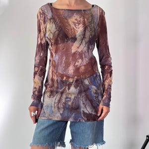 Peut inclure: Un haut à manches longues en maille filet avec un imprimé abstrait de plumes dans les tons marron, bleu et beige, orné de strass argentés sur la poitrine. Le vêtement présente une encolure large et un ourlet droit, porté avec un short en jean bleu effiloché.