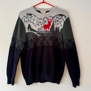 May include: A gray, green, and black sweater with a red deer design. The sweater features a mountain scene and snowflake pattern. The sweater is hanging on a red hanger.