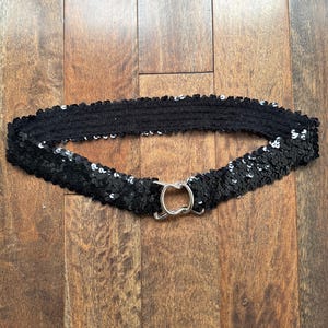 May include: Black sequin belt with a silver-tone metal buckle. The belt is made of a stretchy, elastic material and is covered in small, shiny sequins. The buckle is a simple, circular design.