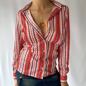 May include: A long-sleeved button-down shirt with a red and white chain pattern. The shirt has a collared neckline and is paired with a dark denim mini skirt. Gold-toned jewelry is also visible.