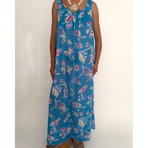 May include: A sleeveless, floor-length blue dress with a colorful abstract pattern. The dress features a scoop neckline and a loose, flowing silhouette. The pattern includes shapes in pink, yellow, and white. The dress is worn with a shell necklace and platform shoes.