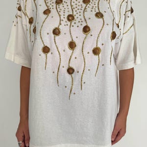 May include: White oversized t-shirt with a gold sequined design of seashells and wavy lines.