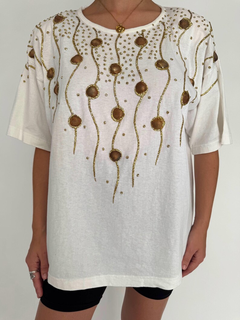 May include: White oversized t-shirt with a gold sequined design of seashells and wavy lines.