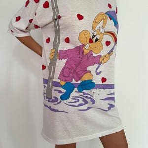 May include: White t-shirt with a cartoon rabbit wearing a pink raincoat and holding an umbrella. The rabbit is standing in a puddle of water with a streetlight in the background. The shirt is covered in red hearts.