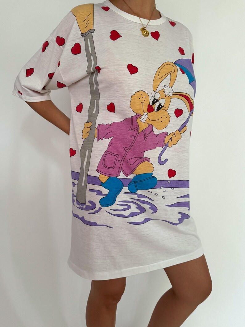 May include: White t-shirt with a cartoon rabbit wearing a pink raincoat and holding an umbrella. The rabbit is standing in a puddle of water with a streetlight in the background. The shirt is covered in red hearts.