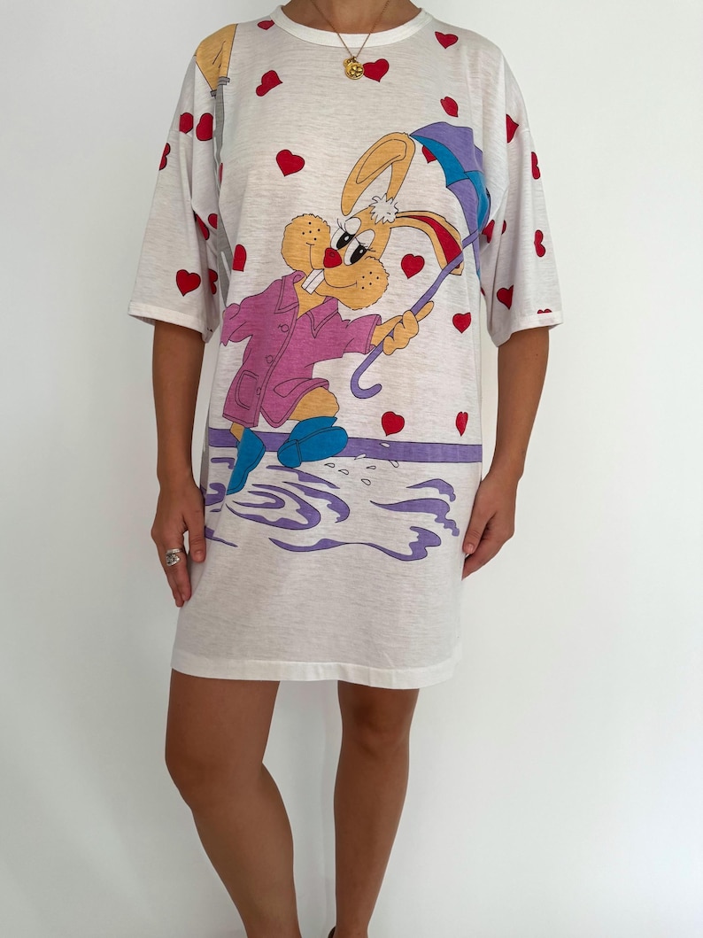 May include: White t-shirt dress with a cartoon bunny wearing a pink raincoat and holding an umbrella. The bunny is surrounded by red hearts.