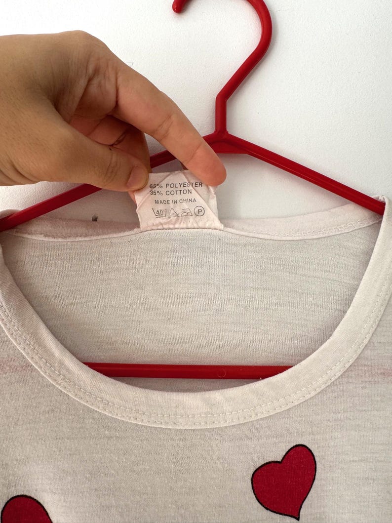 May include: White t-shirt with a red heart graphic print. The tag says "65% Polyester 35% Cotton Made in China".