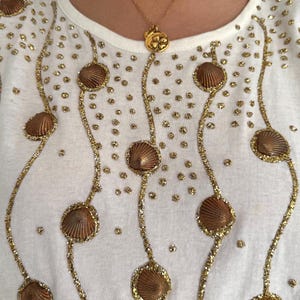 May include: White t-shirt with gold glitter and brown seashell embellishments. The shirt has a round neckline and short sleeves.