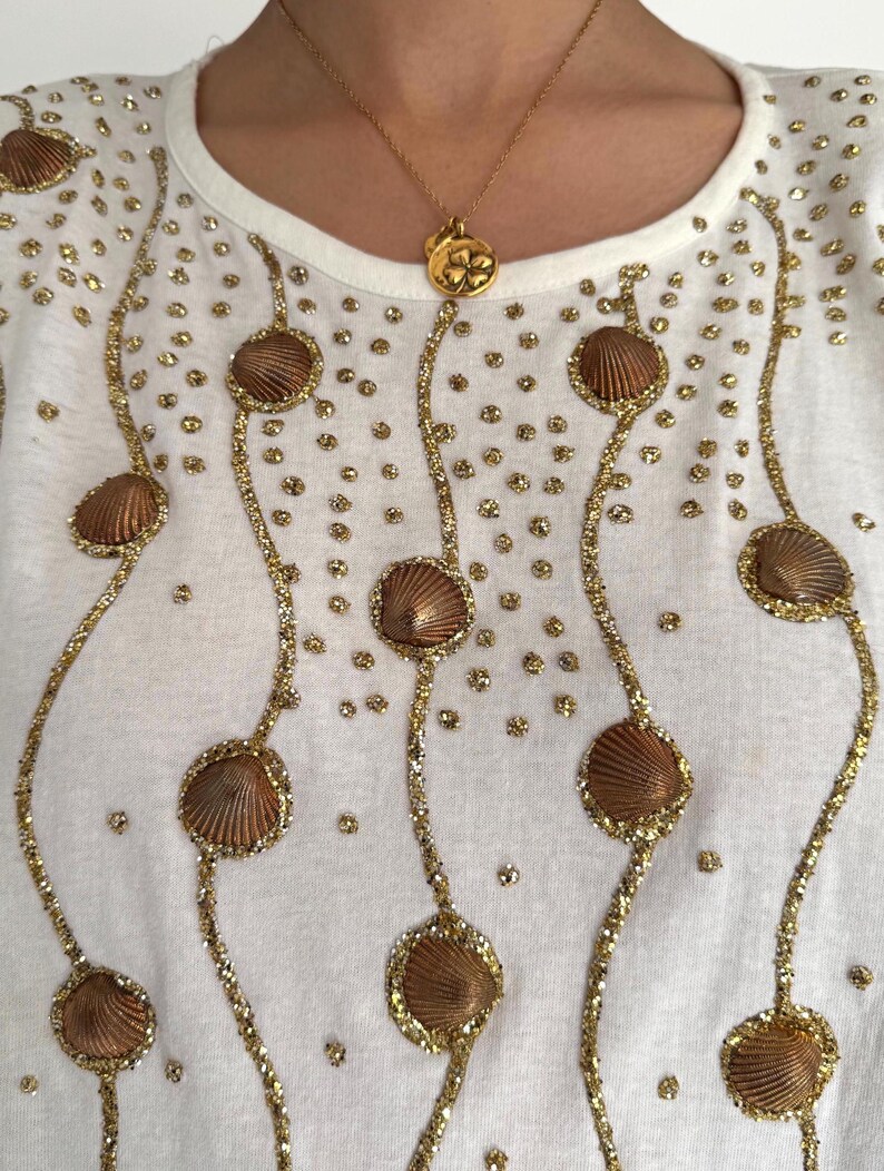 May include: White t-shirt with gold glitter and brown seashell embellishments. The shirt has a round neckline and short sleeves.