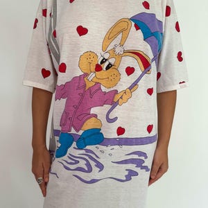 May include: White t-shirt dress with a cartoon bunny wearing a purple raincoat and holding a blue umbrella. The bunny is surrounded by red hearts.