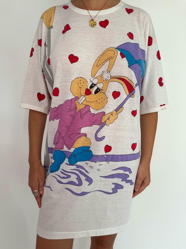 May include: White t-shirt dress with a cartoon bunny wearing a purple raincoat and holding a blue umbrella. The bunny is surrounded by red hearts.
