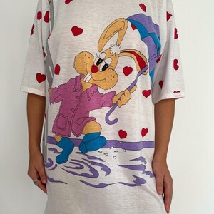 May include: White t-shirt dress with a cartoon bunny wearing a pink coat and holding an umbrella. The bunny is surrounded by red hearts and purple puddles.