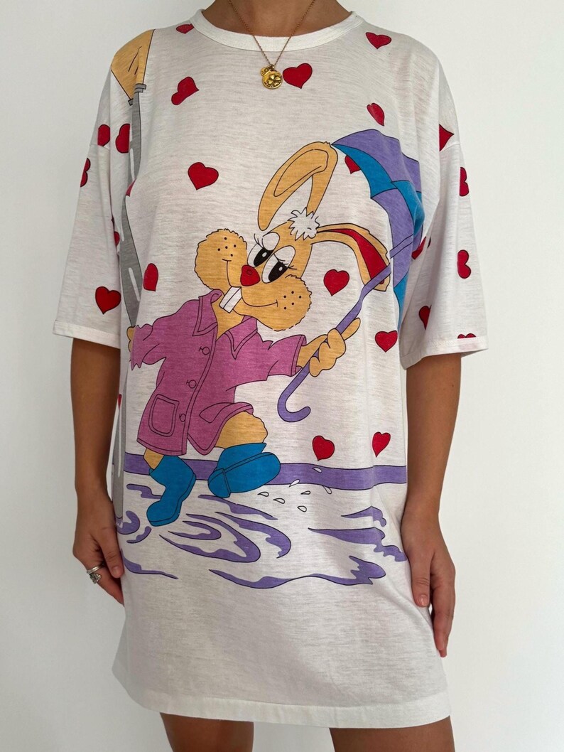May include: White t-shirt dress with a cartoon bunny wearing a pink coat and holding an umbrella. The bunny is surrounded by red hearts and purple puddles.