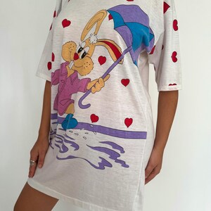 May include: White t-shirt dress with a cartoon bunny holding an umbrella. The bunny is wearing a pink jacket and blue shoes. The background is covered in red hearts.