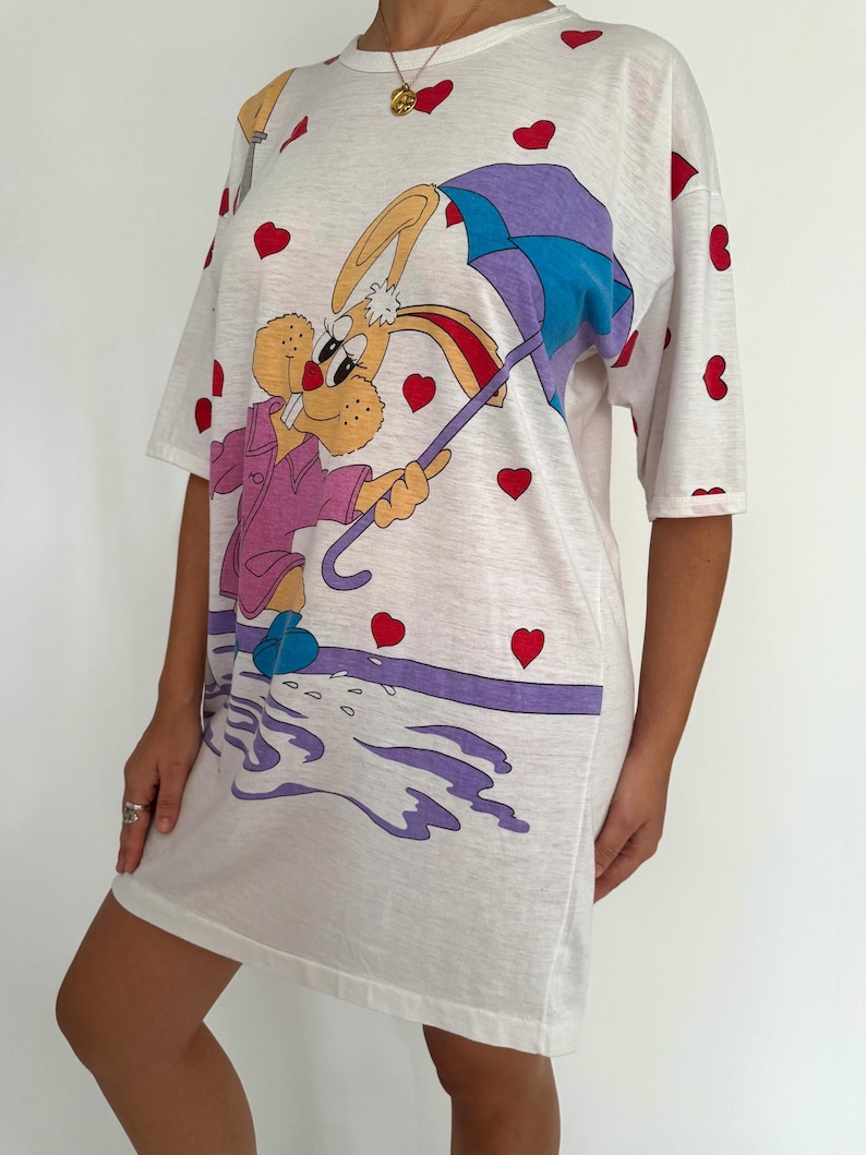May include: White t-shirt dress with a cartoon bunny holding an umbrella. The bunny is wearing a pink jacket and blue shoes. The background is covered in red hearts.