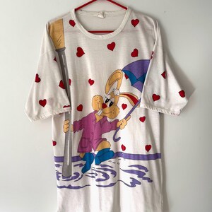 May include: White t-shirt with a cartoon rabbit wearing a purple raincoat and holding an umbrella. The rabbit is standing in a puddle of water with a streetlight in the background. The shirt is covered in red hearts.