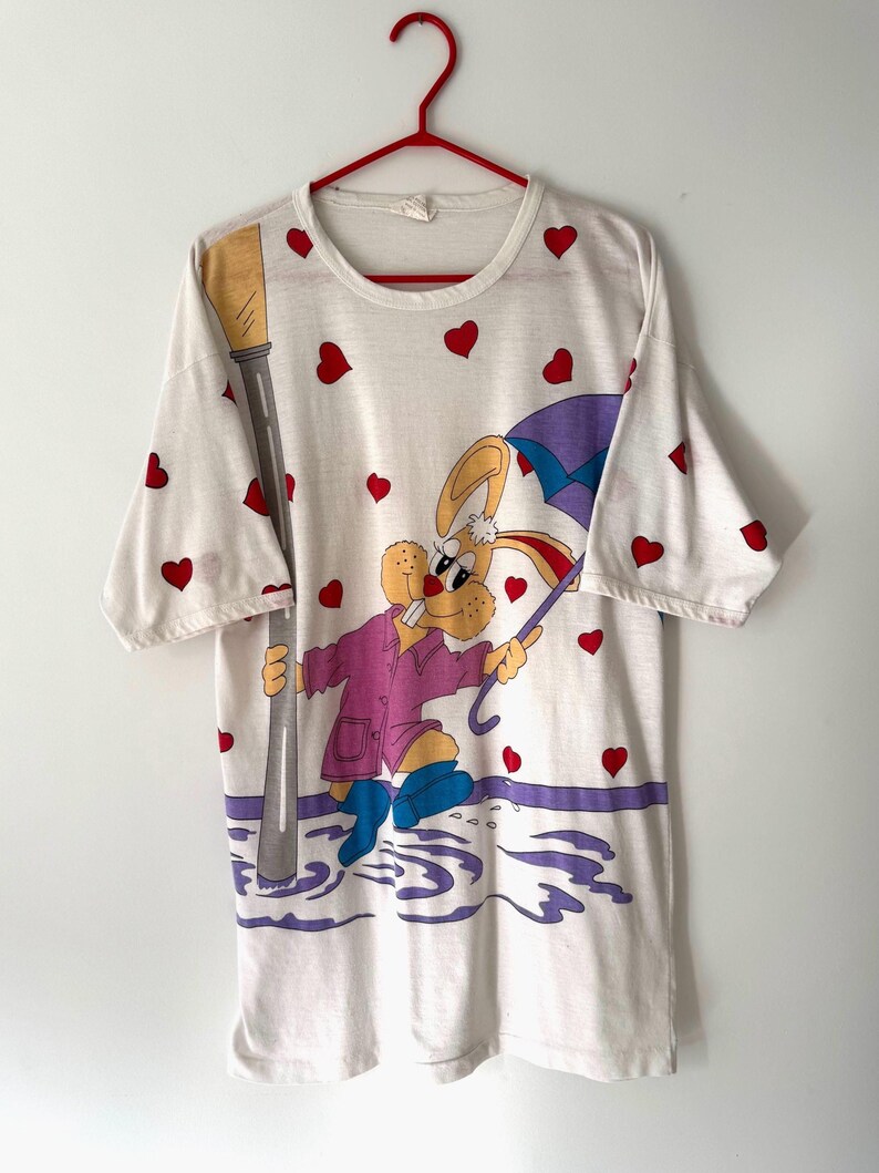 May include: White t-shirt with a cartoon rabbit wearing a purple raincoat and holding an umbrella. The rabbit is standing in a puddle of water with a streetlight in the background. The shirt is covered in red hearts.