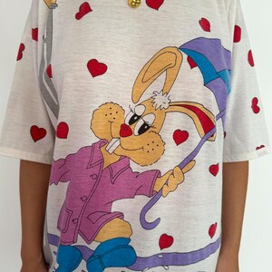 May include: White t-shirt with a cartoon bunny wearing a purple raincoat and holding an umbrella. The bunny is walking through a puddle of water. The shirt is covered in red hearts.