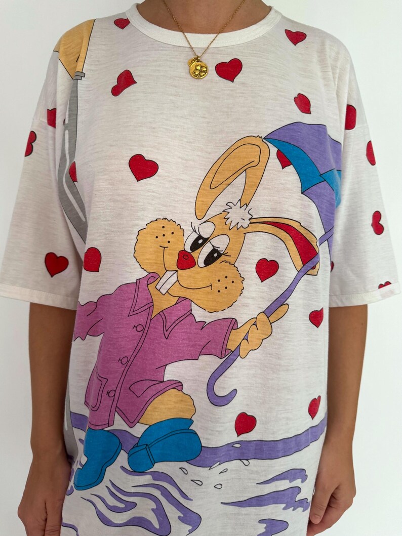 May include: White t-shirt with a cartoon bunny wearing a purple raincoat and holding an umbrella. The bunny is walking through a puddle of water. The shirt is covered in red hearts.